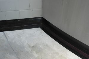 Een detailopname van de hoek van een kelder waar de vloer en de wand samenkomen. Er is een vloeibare rubber aangebracht langs de randen om de kelder dicht te maken en lekkage te voorkomen. De afdichting bedekt de voeg tussen de betonnen vloer en de betegelde wand, en loopt door langs de onbetegelde muur.